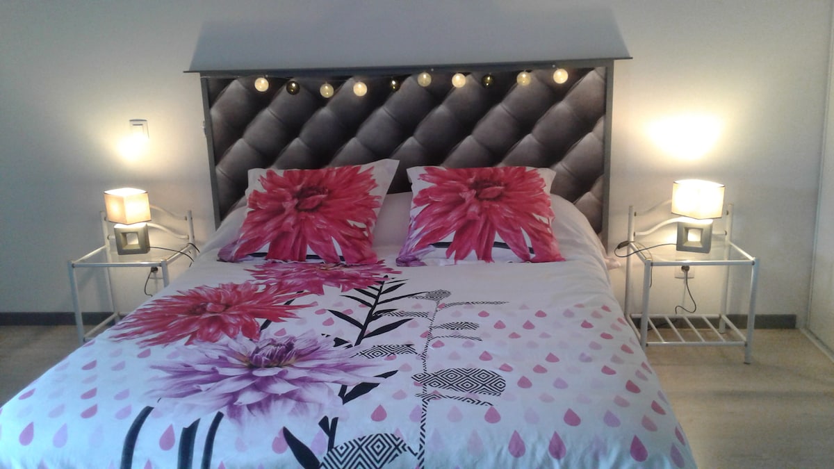 A double bed features a decorative floral bedding set, complemented by contrasting flower-patterned pillows. Soft lighting is provided by two bedside lamps placed on minimalistic white nightstands, enhancing the calming ambiance of the space.