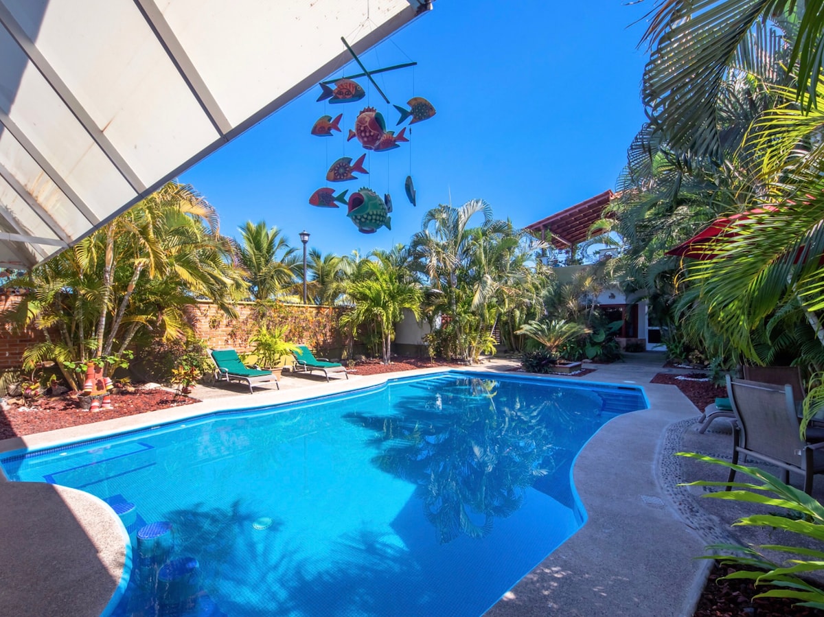 A tranquil pool is framed by lush palm trees and vibrant flowers, creating a serene outdoor space. Two lounge chairs are positioned by the water, while colorful fish decorations dangle overhead, adding a playful touch to the atmosphere.
