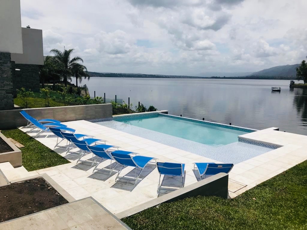 A shared swimming pool is situated beside a tranquil lake, framed by lush greenery. Rows of blue lounge chairs line the poolside, providing ample space for relaxation. The calm water reflects the surrounding sky, contributing to the serene environment.