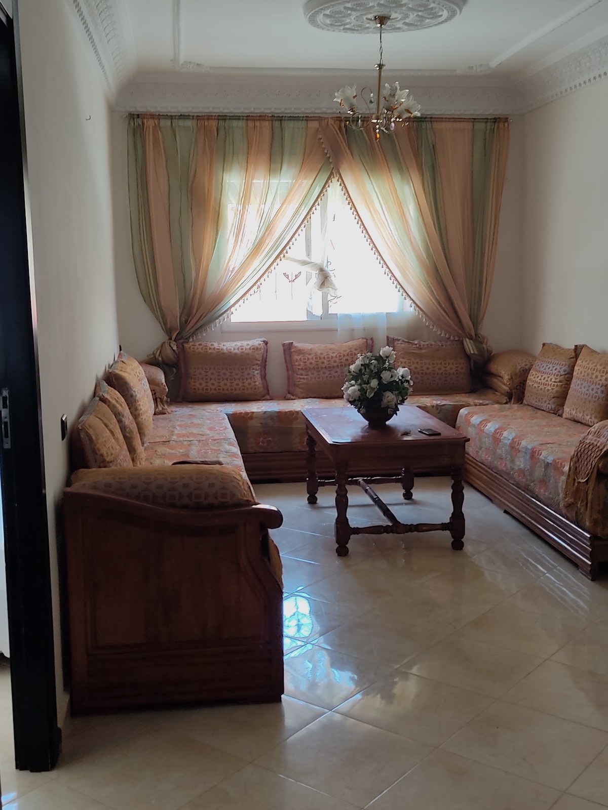 A cozy living area features a set of upholstered sofas arranged around a wooden coffee table. Natural light enters through a window dressed with flowing curtains. A vase of flowers adorns the table, adding a touch of liveliness to the warm-toned decor.