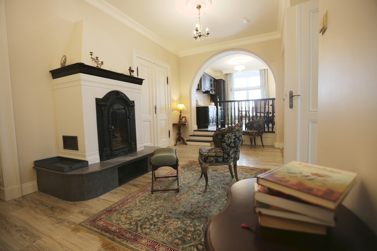 The inviting living area features a decorative fireplace with an ornate black mantel. A plush chair sits beside a small green chair on a patterned rug. Light enters through an arched doorway leading to a spacious area, showcasing a blend of classic and contemporary elements.