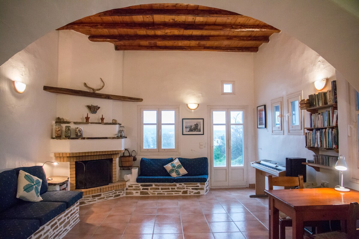 A cozy living area features beamed ceilings and terracotta tile flooring. Built-in sofas flank a traditional fireplace, while a dining table and chairs are positioned nearby. A Yamaha piano and shelves of books add to the welcoming ambiance. Natural light streams in through the windows, revealing scenic views.