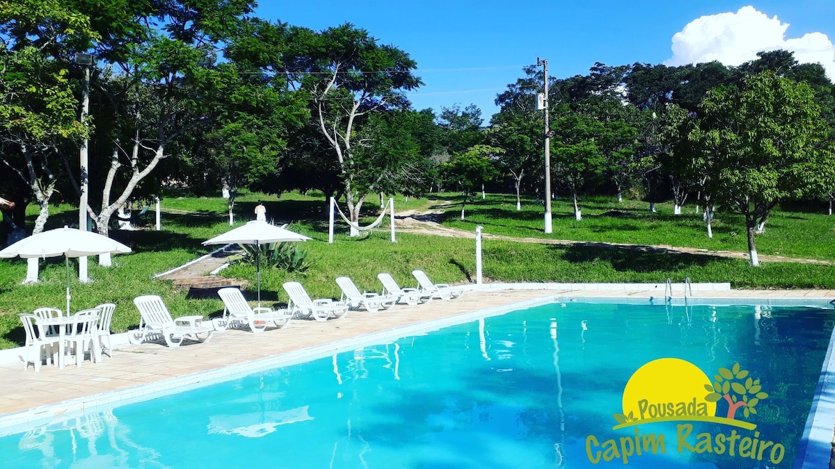 A clear blue pool is surrounded by sun loungers and umbrellas, set against a backdrop of lush greenery. The well-maintained area allows for relaxation while enjoying the natural environment. Bright skies and trees create a tranquil outdoor setting.