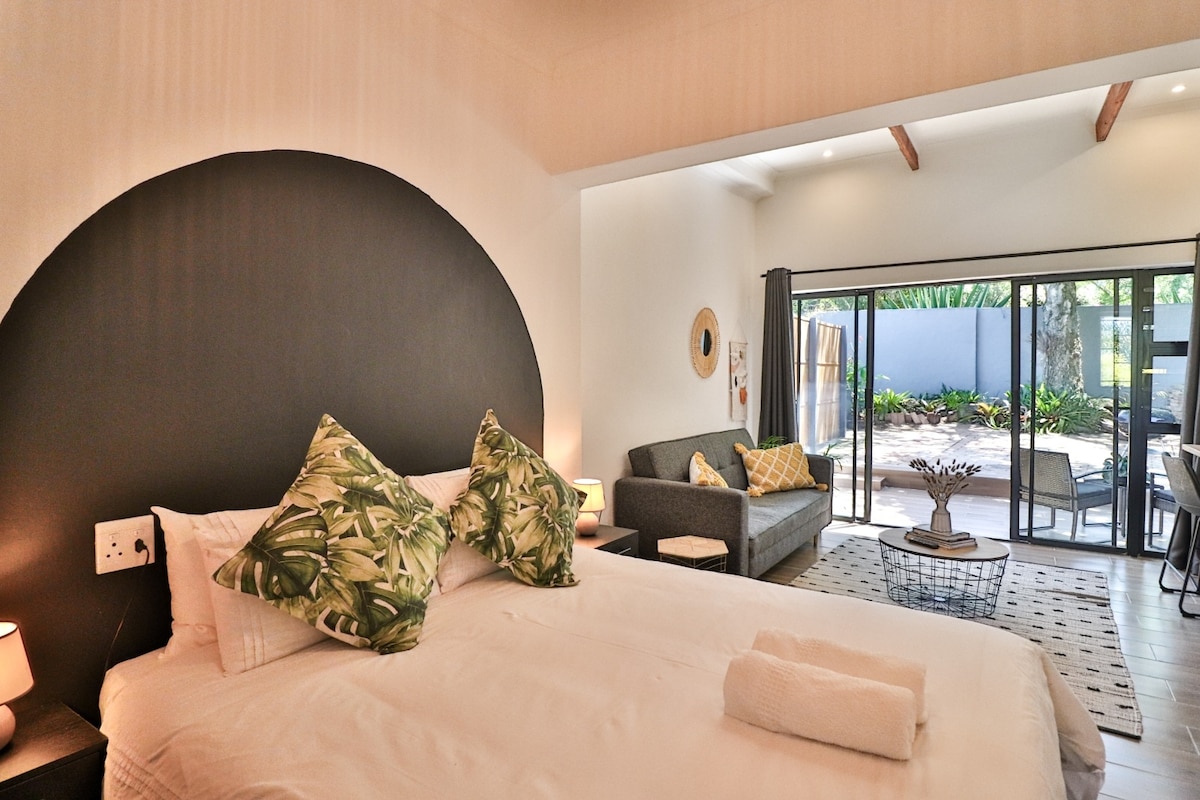 A well-lit sleeping area features a spacious bed adorned with patterned pillows. A cozy seating area includes a sofa, with large glass doors providing access to an outdoor space. The design blends natural elements, creating a harmonious atmosphere.