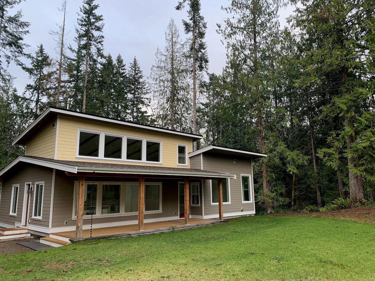 A modern single-level home is nestled in a forested setting, featuring large windows that allow natural light to fill the space. The exterior showcases a spacious covered deck and a green lawn area, surrounded by trees that enhance the tranquil atmosphere.