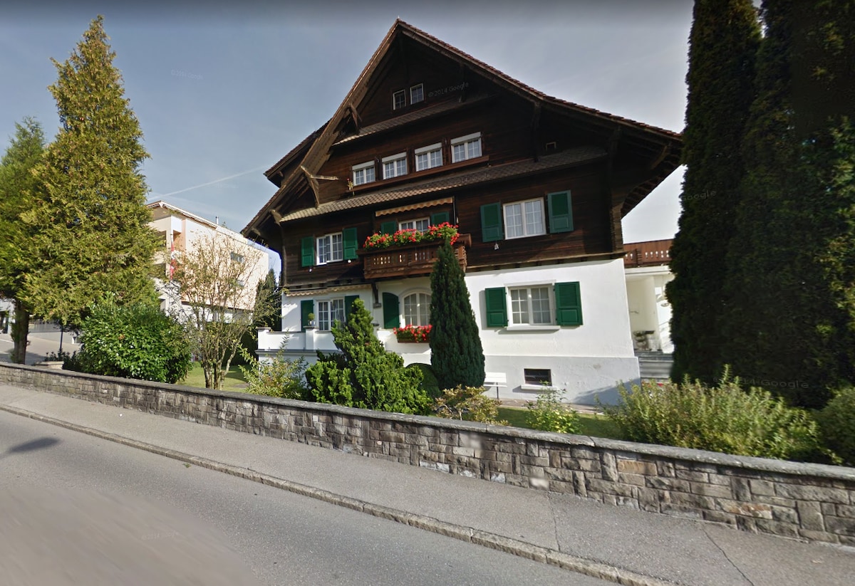 The exterior of a charming three-story house is shown, featuring dark wooden siding and green shutters. Flower boxes adorn the balconies, and neatly trimmed shrubs frame the entrance. A stonewall delineates the property from the road, enhancing the quaint character of the building.