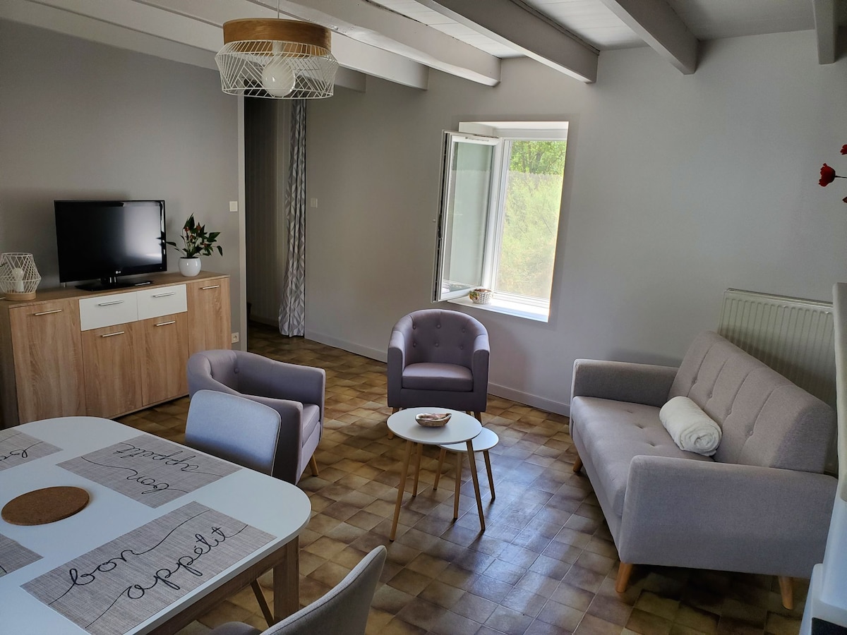 A welcoming living area features light-colored walls and a combination of modern furnishings. A small round table is set with placemats, while a comfortable sofa and two armchairs provide seating. Natural light enters through an open window, illuminating the space.