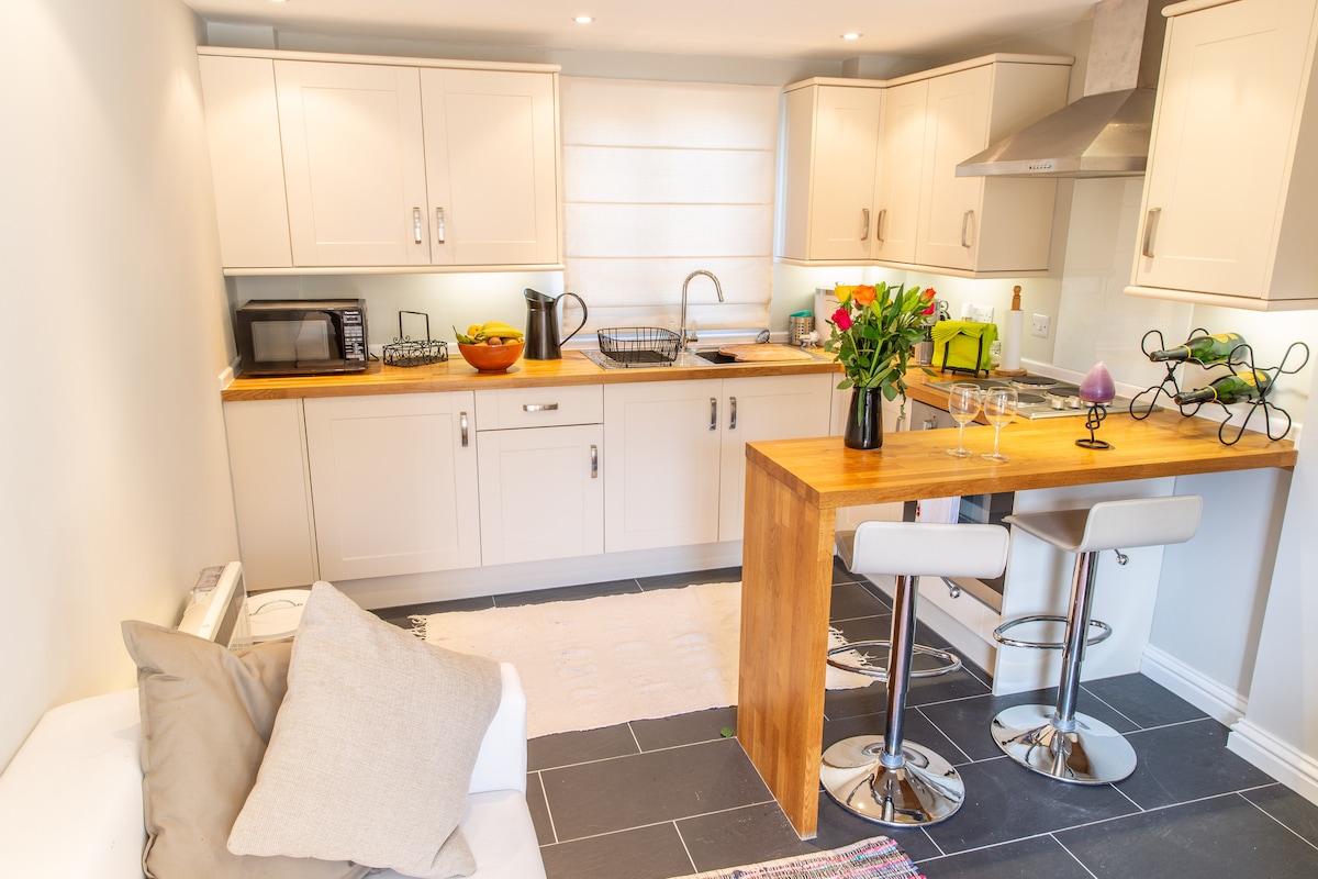 A modern kitchen area features light-colored cabinetry and a wooden countertop, providing ample workspace. Stainless steel appliances are included, alongside a dining table with two high-backed stools. Fresh flowers are arranged on the table, and light floods in through a nearby window.