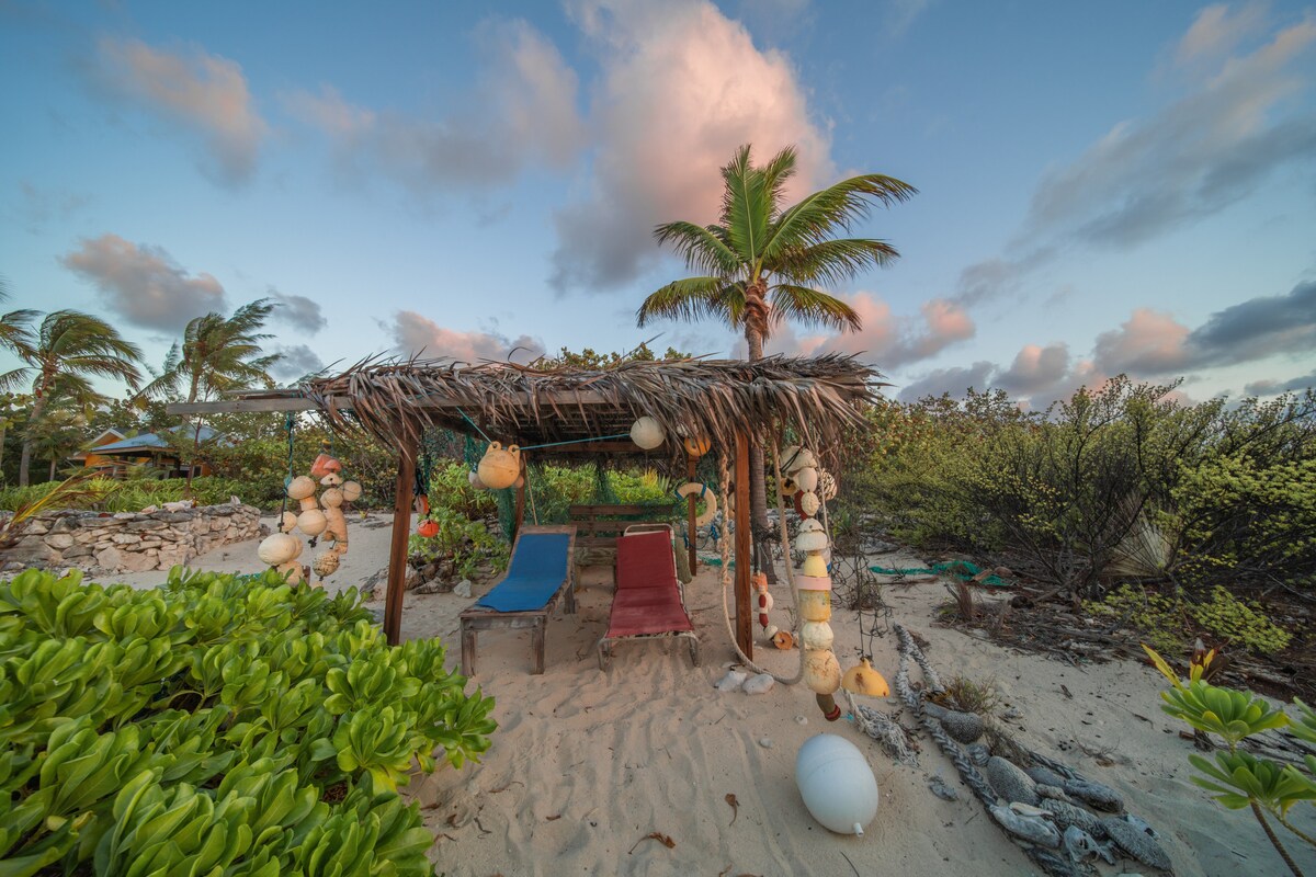 A shaded cabana is nestled among sandy ground and tropical foliage. Two lounge chairs, one blue and one red, are positioned beneath a thatched roof adorned with natural decorations. Nearby palm trees sway gently, creating a relaxed outdoor environment suitable for leisure.