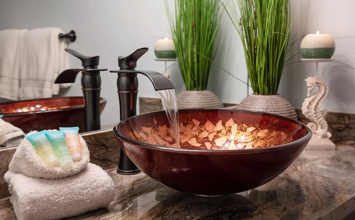 The bathroom features a vibrant bowl sink with a gently flowing faucet, complemented by a granite countertop. Soft towels are neatly arranged beside the sink, alongside a selection of toiletries in various colors. Decorative plants provide a fresh touch, while candles create a calming ambiance.