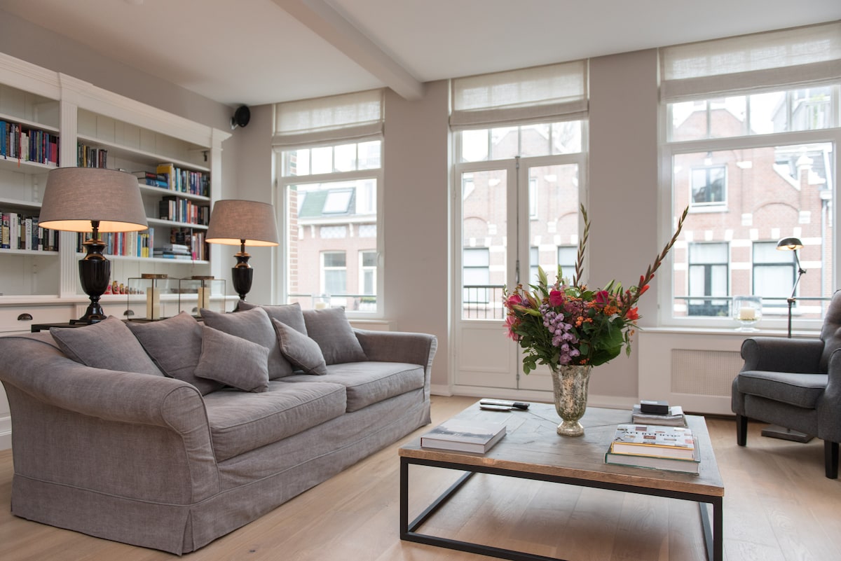 A spacious living room features a comfortable grey sofa adorned with cushions. A coffee table sits in front, displaying books and a floral arrangement. Natural light filters through large windows, highlighting the bright and airy atmosphere. Bookcases line one wall, adding functional elegance.