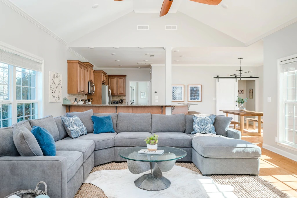 A spacious living area features a large, L-shaped gray sectional sofa adorned with blue accent pillows. A circular glass coffee table is centered on a textured area rug, while natural light floods the room through multiple windows, highlighting the open layout that connects to the kitchen.
