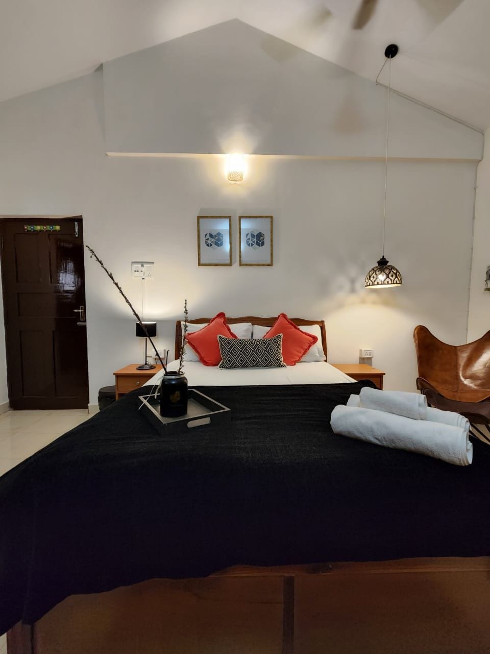 A spacious sleeping area is highlighted by a large bed featuring decorative pillows and a neatly folded towel. A bedside table holds a tray with items, and a unique light fixture adds character. Natural light enters the room from above, creating an inviting ambiance.