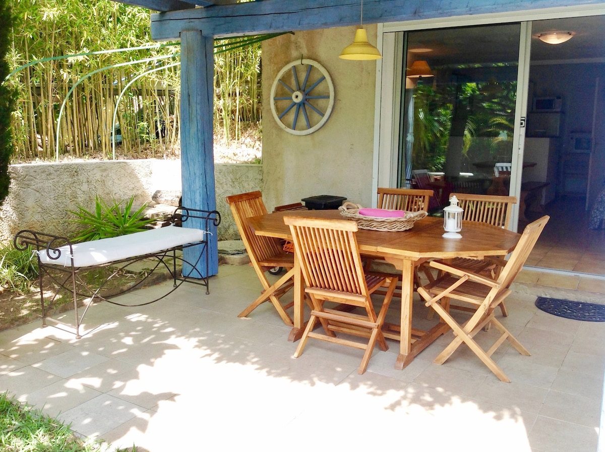 An outdoor dining area is presented, featuring a wooden table surrounded by six folding chairs. A bench lies nearby, and greenery provides a natural backdrop. Soft sunlight illuminates the space, while a decorative wheel adds a charming touch to the wall.
