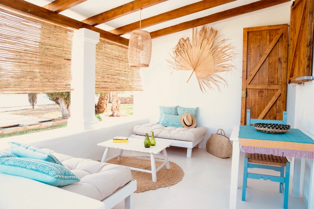 A light and airy living space features two comfortable sofas arranged around a low coffee table. Natural light floods in through woven shades, illuminating the white walls and rustic wooden beams overhead. A decorative palm leaf hangs against the wall, complementing the tranquil atmosphere.