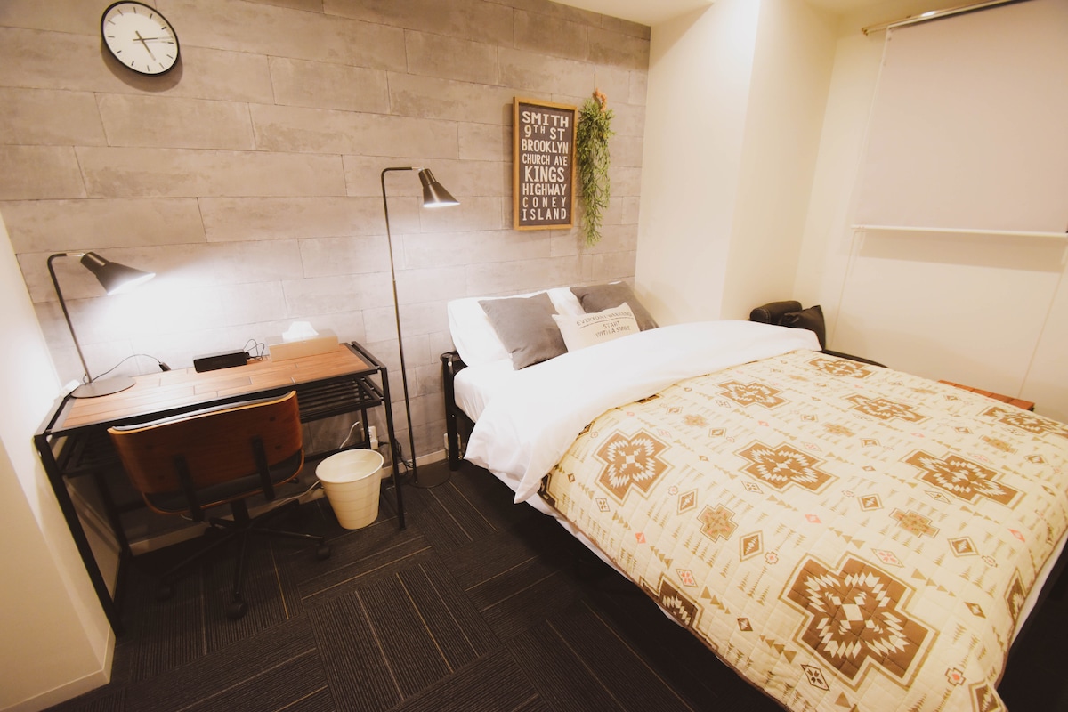 A well-lit room features a spacious bed with a patterned blanket. A desk with a chair is positioned nearby, complemented by a table lamp. A wall clock and decorative wall art add character, alongside a small plant hanging above the bed.