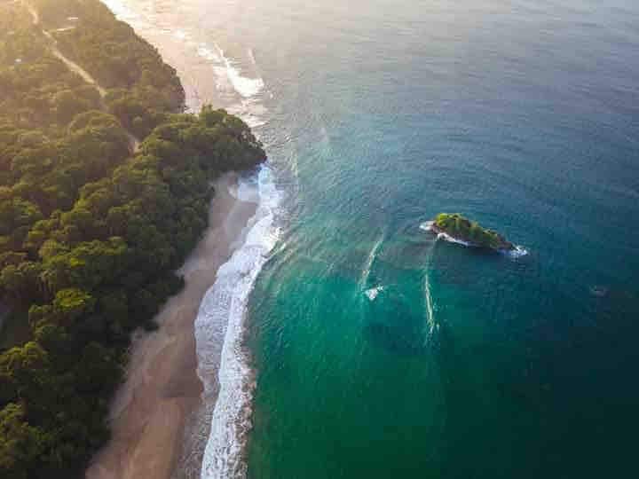 Aerial view captures a stretch of sandy beach bordered by lush greenery. Gentle waves lapping at the shore meet turquoise waters, with a small island visible offshore. The serene landscape showcases the natural beauty of the Caribbean coastline.