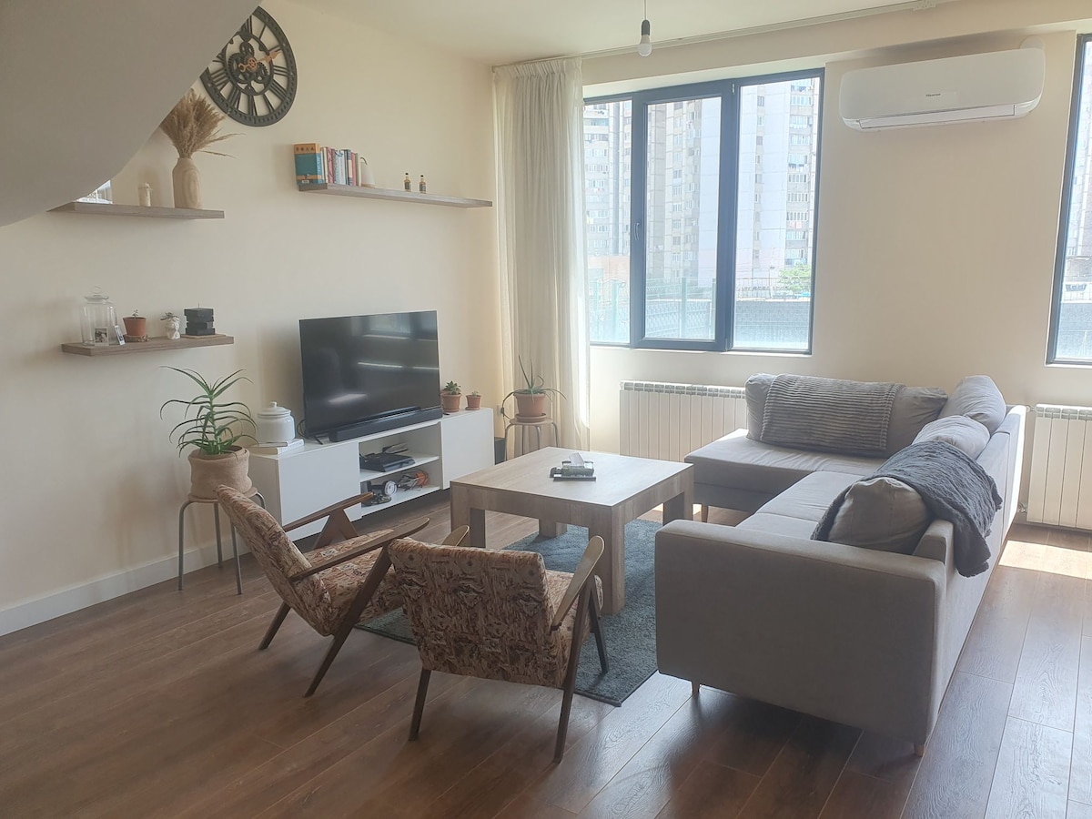 A modern living area is showcased, featuring a sofa arranged with decorative pillows and a plush armchair. A coffee table sits in front of the sofa, accompanied by a television mounted on the wall. Natural light enters through large windows, highlighting the warm wooden flooring.