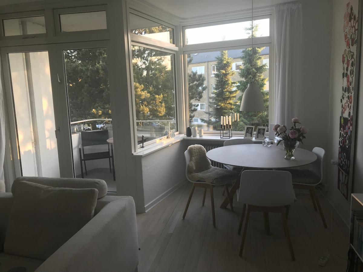 A dining area is presented with a round table surrounded by white and wooden chairs. Large windows allow natural light to fill the space, while a vase of flowers enhances the setting. A cozy sofa is positioned nearby, contributing to a relaxed atmosphere.