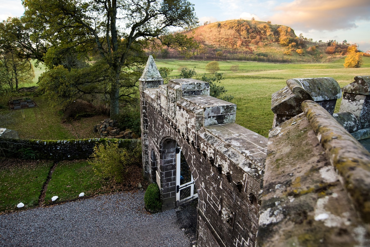 A view showcases the historic gatehouse with its stone architecture, framed by expansive green fields and gentle hills in the distance. The landscape reflects a natural setting, accented by the warm colors of autumn foliage and a calm sky.