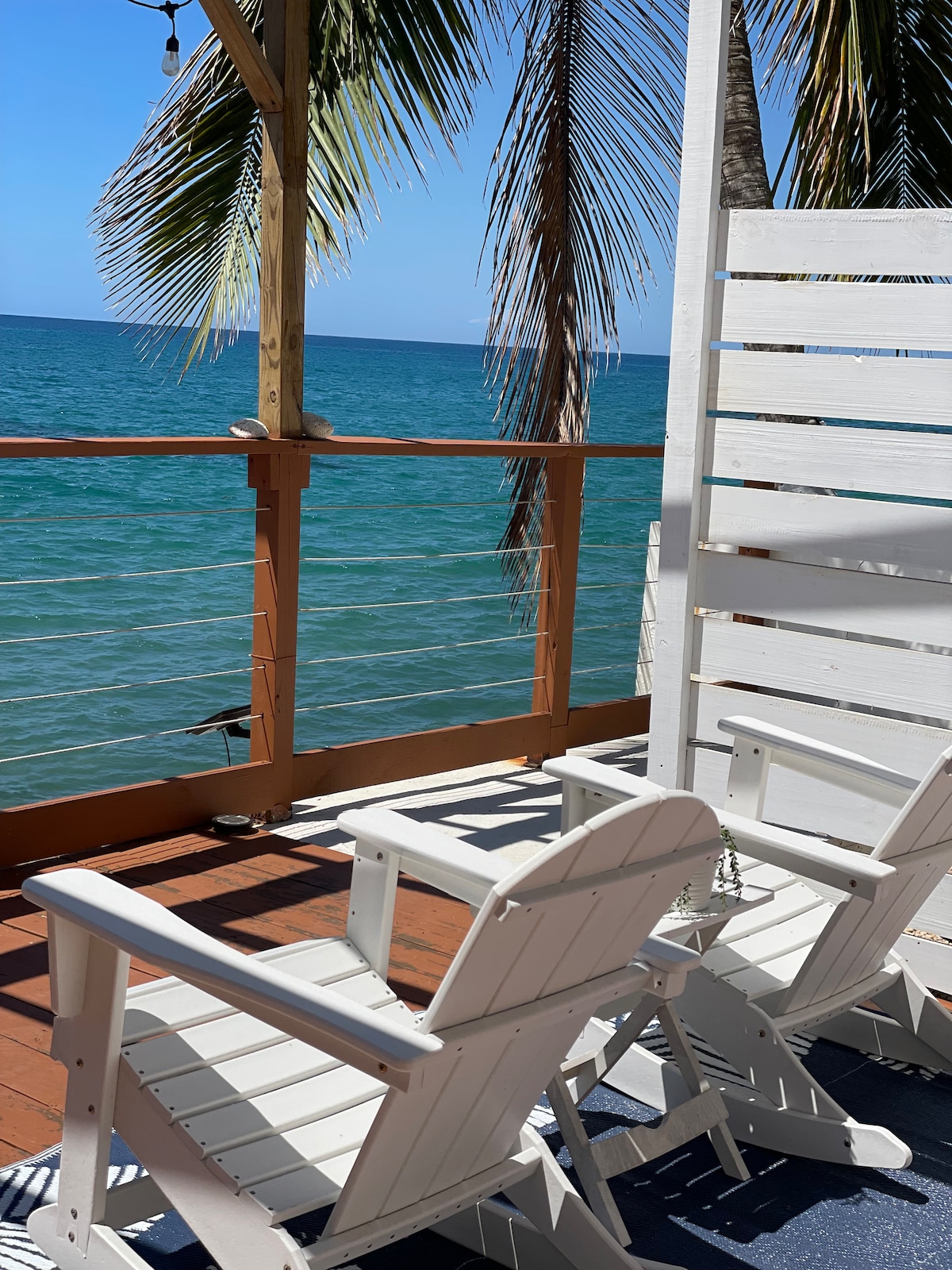 Two white outdoor chairs are positioned on a wooden deck, facing the ocean. Palm fronds gently sway above, framing a clear blue view of the water. The railing provides a sense of openness while ensuring privacy, creating a serene outdoor space for relaxation.