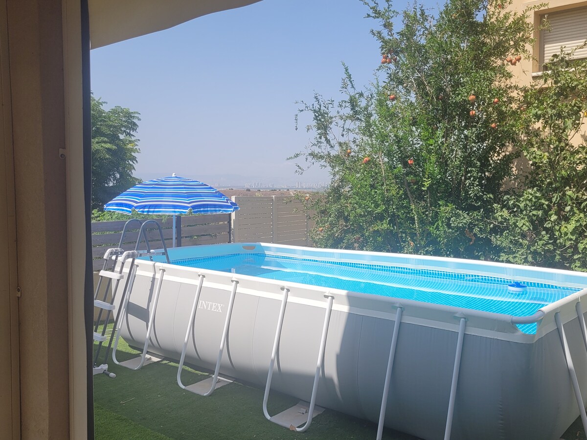 An above-ground pool is situated in a grassy area, framed by lush greenery and a blue-and-white striped umbrella. A ladder leans against the pool's side, providing access. The sky appears clear, emphasizing a tranquil outdoor setting.