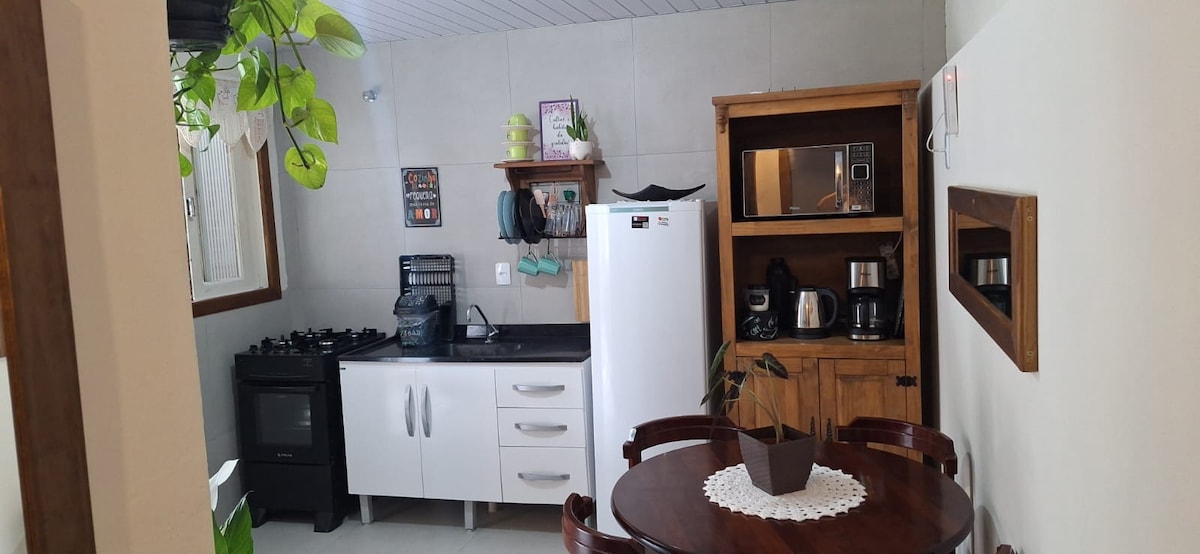 A compact kitchen area features a modern white refrigerator, a gas stove, and a wooden table set for four. Essential appliances, including a microwave and coffee maker, are neatly arranged on the countertop, complemented by a decorative plant adding a touch of greenery.