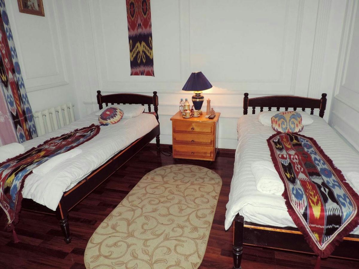 A room with two single beds arranged side by side, each adorned with colorful blankets. A bedside table with a lamp and decorative items sits between the beds. A soft area rug adds warmth to the wooden floor, and patterned wall hangings enhance the decor.