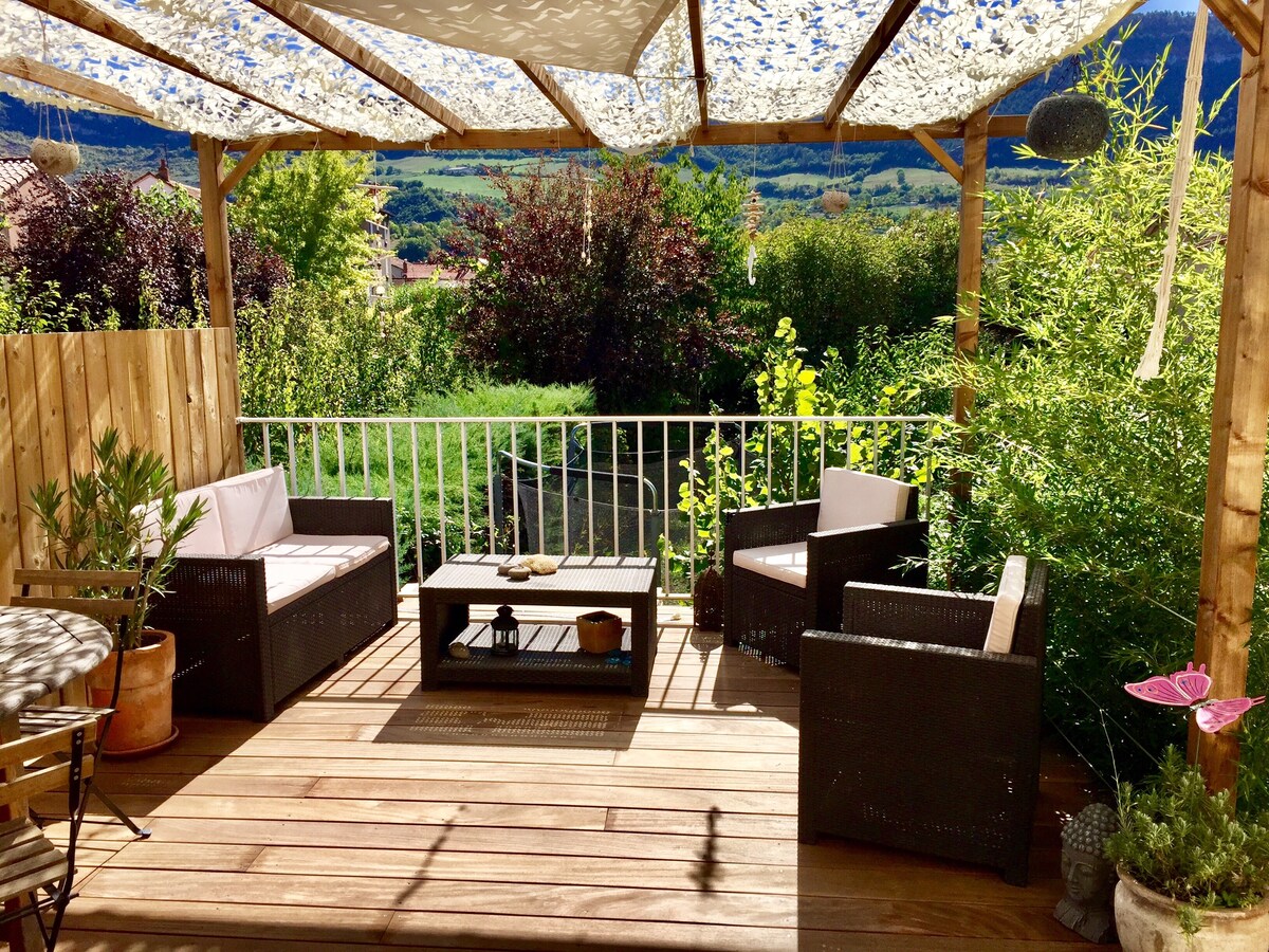 A wooden terrace is presented, featuring a pergola adorned with fabric drapes. Two outdoor chairs and a low coffee table create a comfortable seating area, surrounded by greenery and a tranquil view. The flooring consists of smooth wooden planks, enhancing the inviting outdoor space.