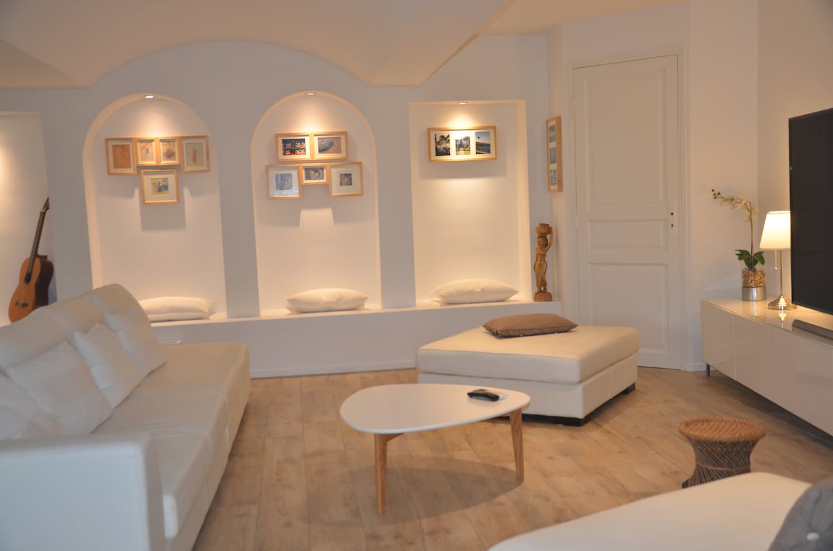 A light-filled living area is highlighted by white walls and a wooden floor. Comfortable seating options are arranged, including a cream-colored sofa and an ottoman. Decorative alcoves display framed photos, and soft pillows are positioned on the ledges, offering a cozy, inviting space.