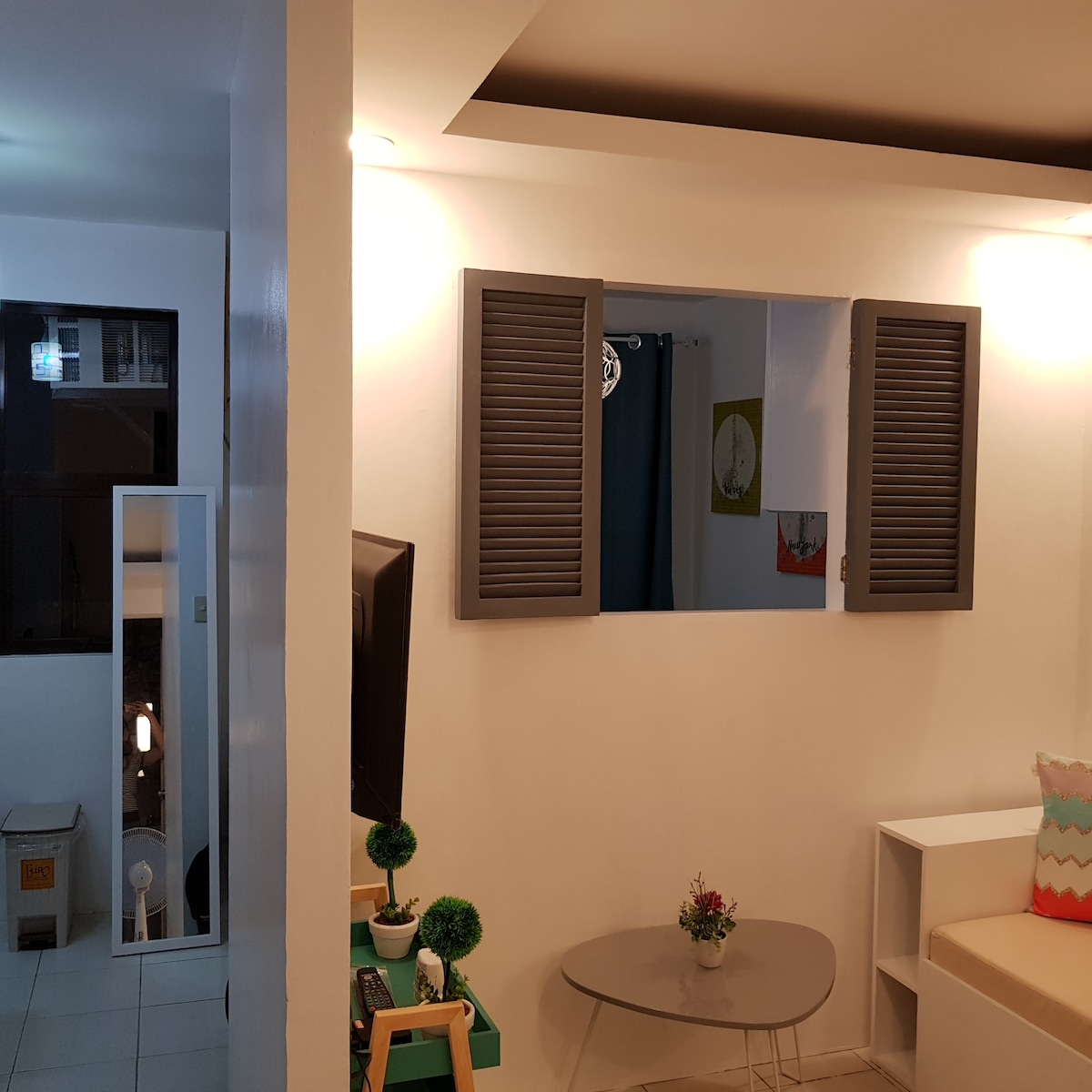 An open-plan living area features a cozy seating arrangement with a light-colored sofa and a round table. Window shutters frame a view of the outside, while a small decorative plant sits on the table. A television is mounted on the opposite wall.
