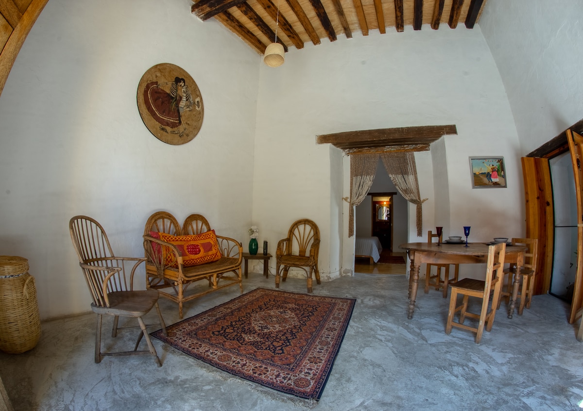 A spacious living area features wooden beams overhead and a traditional woven rug on a stone floor. A variety of wooden seating options are present, along with a dining table. Soft light enters through a doorway leading to the bedroom area, enhancing the serene ambiance.