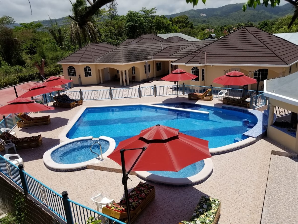An outdoor area features a large swimming pool surrounded by sunbeds and red umbrellas. A hot tub is positioned at one end of the pool. Lush greenery and distant mountains create a serene backdrop, enhancing the inviting atmosphere of this relaxing space.