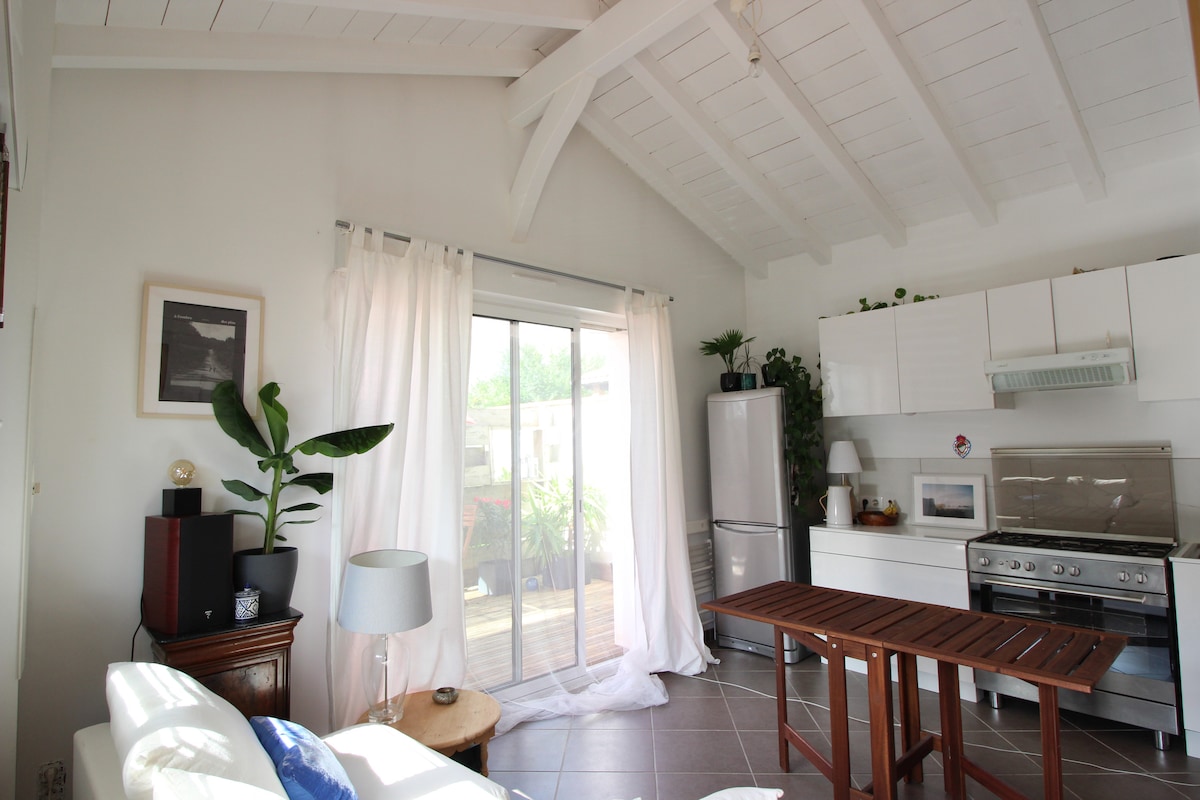 A bright interior space is featured with a vaulted ceiling and light-colored wooden beams. A sliding glass door opens to a terrace, allowing natural light to fill the room. A dining table is positioned centrally, surrounded by modern kitchen elements and indoor plants.
