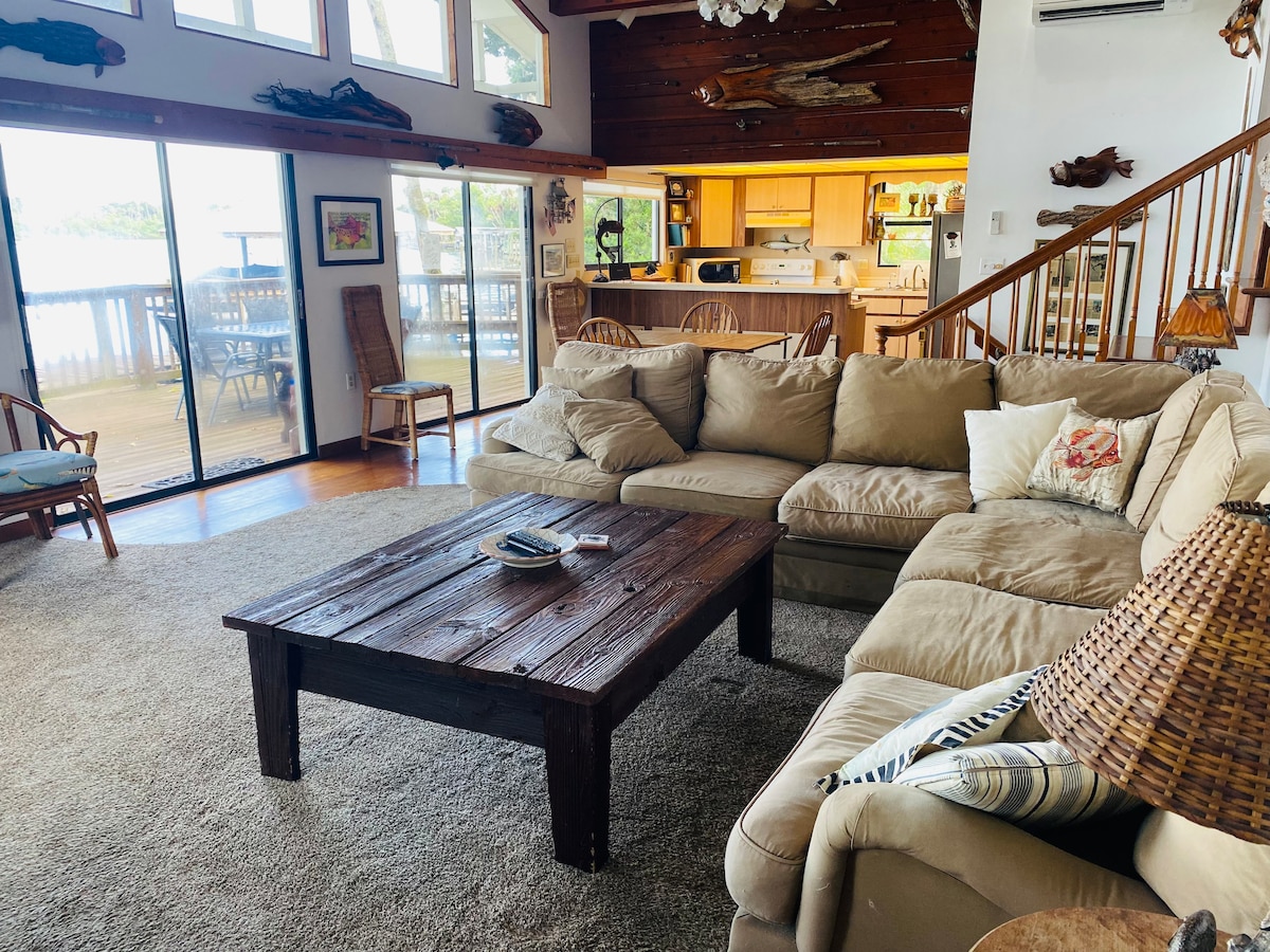 A spacious living area is furnished with a large, comfortable sectional sofa and a rustic wooden coffee table. Sunlight streams in through expansive glass doors, offering views of the outdoors. A light-colored rug covers the floor, complementing the natural wooden accents throughout the space.