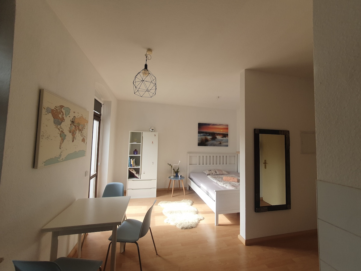 A bright room features a spacious bed with a light-colored frame. A small dining table with two blue chairs is present, along with a white wardrobe. A world map hangs on the wall, and a large mirror reflects the natural light streaming through the window.