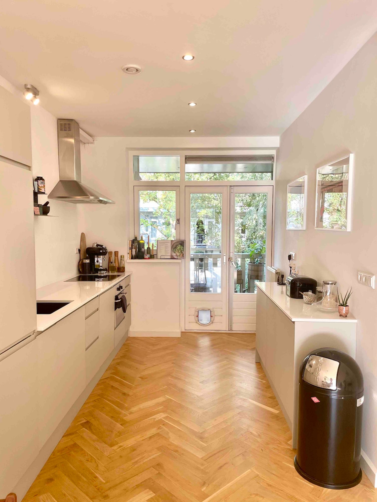 A modern kitchen features light-colored cabinets with a subtle texture, complemented by stainless steel appliances. Large windows provide natural light and a view of greenery outside. A herringbone-patterned wooden floor adds warmth, while a sleek trash can sits discreetly in the corner.