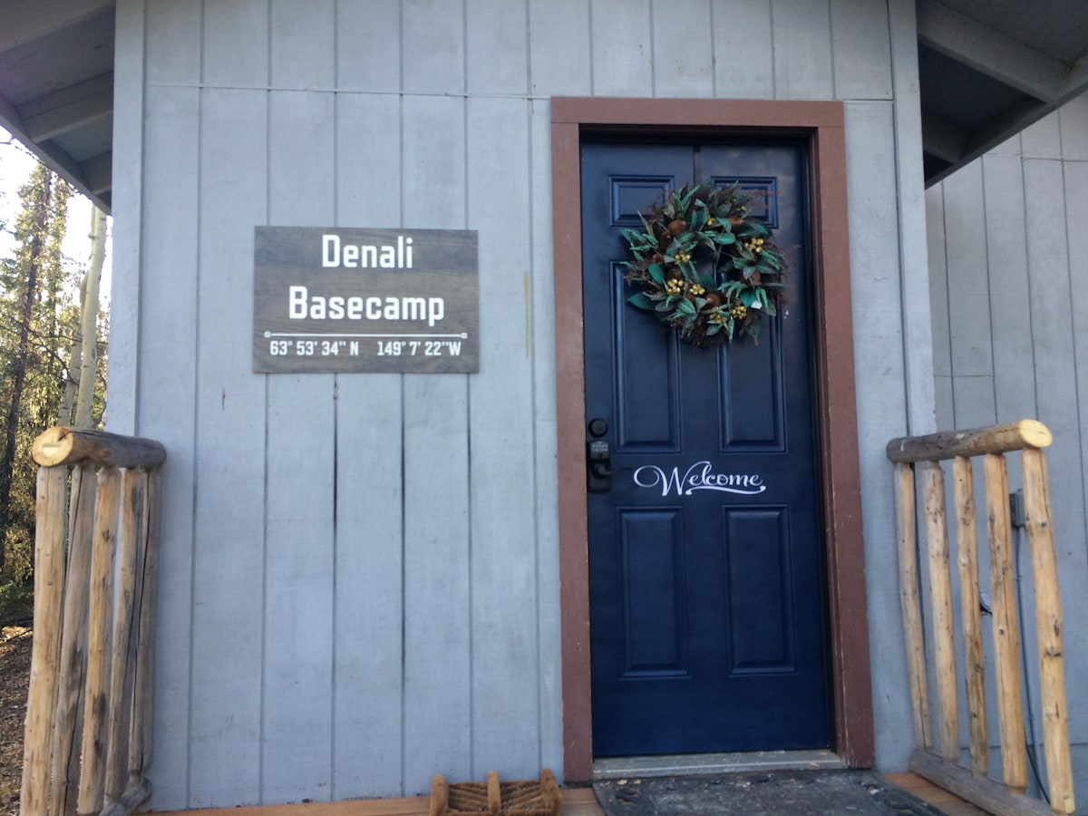 The entrance to the cabin is framed by wooden beams with a welcoming sign displayed on the wall. A dark blue door is adorned with a decorative wreath, inviting guests into the rustic space.