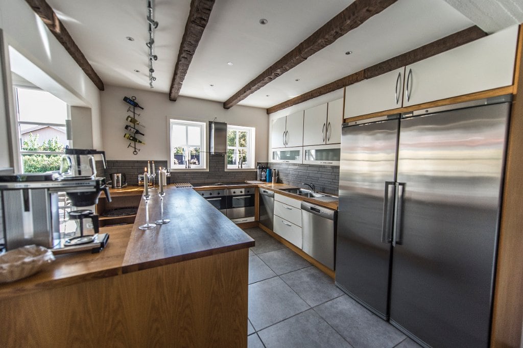 The kitchen features modern appliances and wooden cabinetry. A spacious countertop extends alongside stainless steel refrigerators. Natural light enters through the window, illuminating the tiled floor and creating an open, inviting workspace.