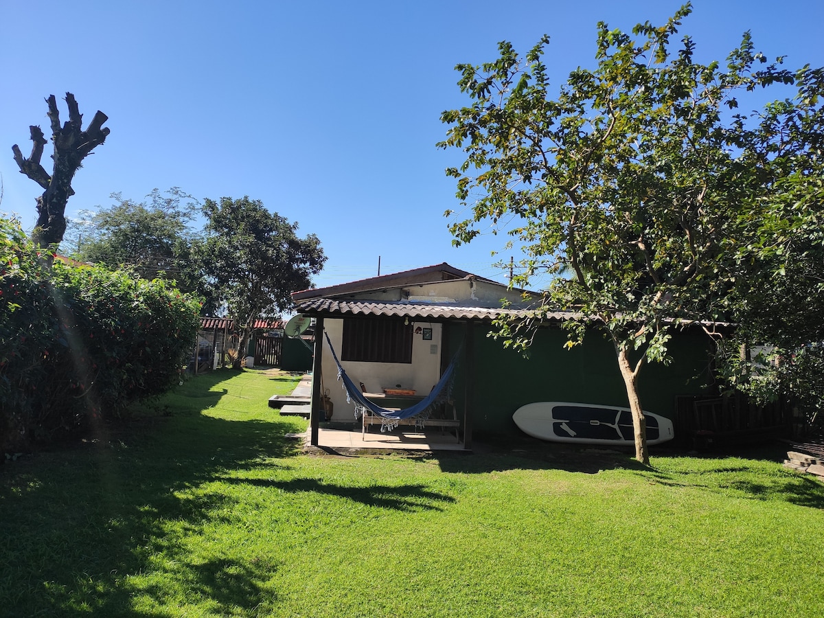 A private quintal is shown, featuring a grassy area surrounded by trees and shrubs. A charming structure with a shaded porch and hammock is visible, alongside a paddleboard leaning against the wall. Sunlight brightens the scene, creating a peaceful outdoor atmosphere.