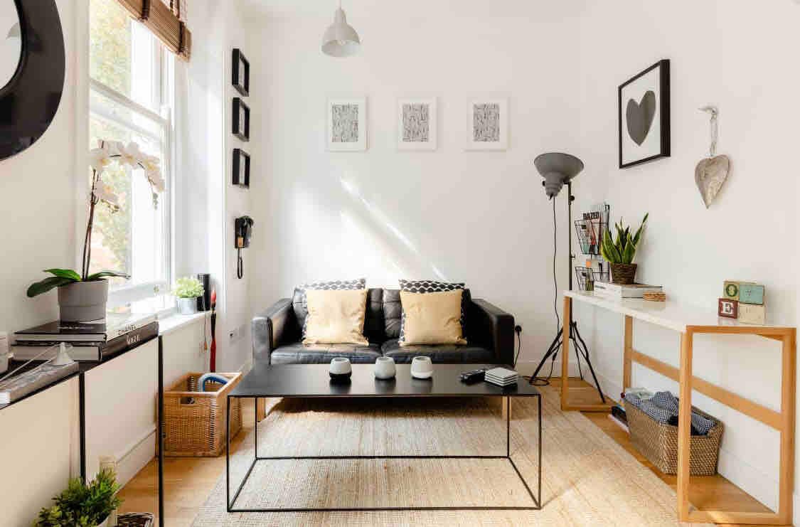 The lounge features a black leather sofa facing a rectangular coffee table. Natural light filters through sash windows, highlighting botanical elements and framed artwork on the walls. A console table with decorative items and plants provides additional interest to the space.