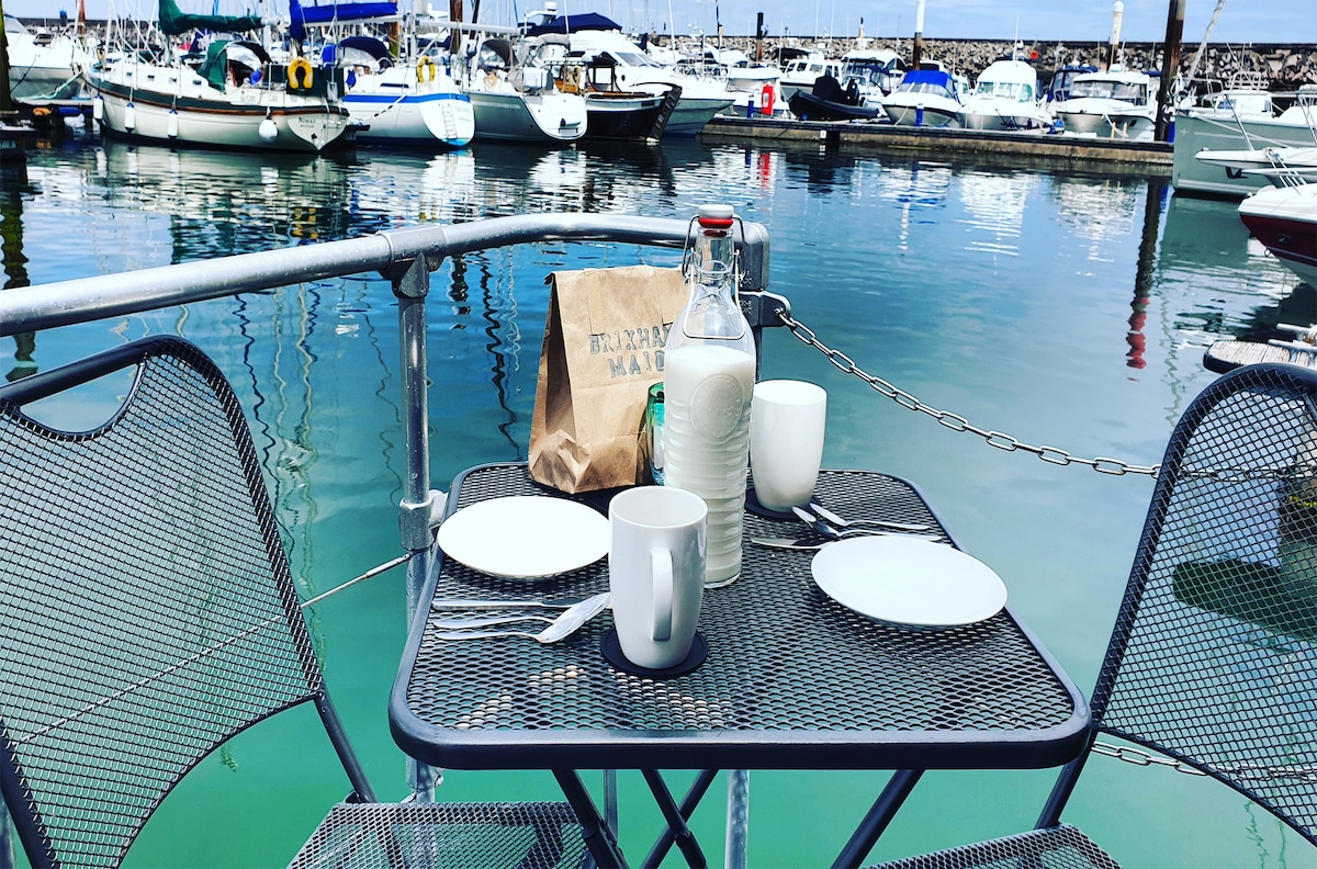 A metal table is set for two on the deck of a boat, featuring two empty plates and cups. A glass bottle filled with water is positioned in the center, with a paper bag resting nearby. The calm marina water reflects the surrounding boats and sky.