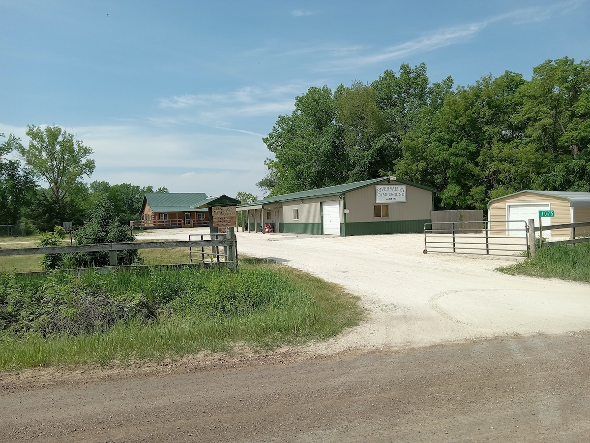 A gravel driveway leads to a spacious property, flanked by greenery and trees. The main building features a sign indicating its use as a rental with a large outdoor area. Additional structures are visible, and ample parking space is available.
