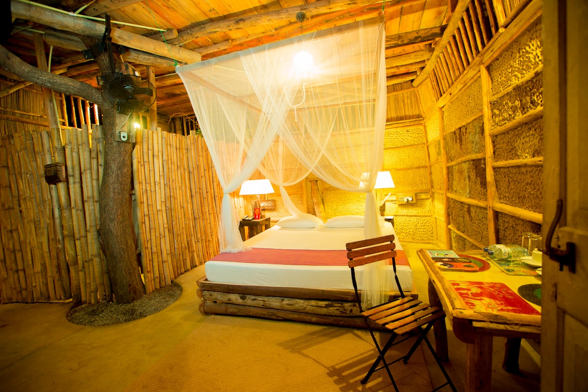 A cozy chalet interior features a spacious bed draped in white mosquito netting. Soft lighting is provided by two bedside lamps. A wooden table and chairs, alongside textured bamboo walls, create a warm and natural atmosphere. A tree trunk is integrated into the room's design.