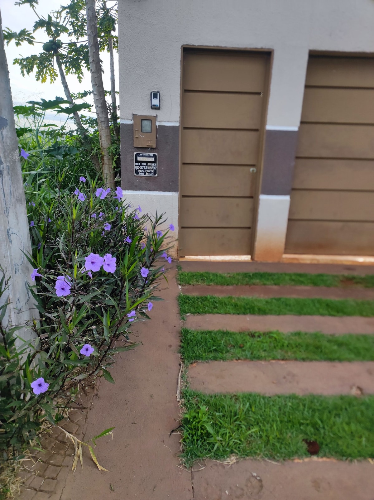 A neat entrance area is depicted, featuring a brown garage door set against a light-colored wall. Lush green grass lines the path, complemented by purple flowers growing along the side. The scene is framed by trees that provide a touch of greenery.