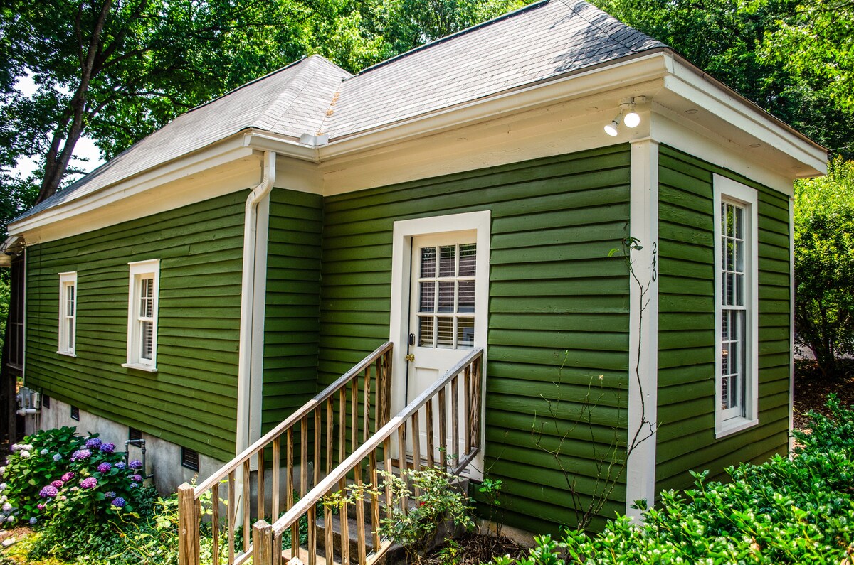 A charming green cottage exterior is captured amidst lush trees. The entrance features a wooden railing leading up to a welcoming door, flanked by multiple windows. The vibrant colors of flowering bushes at the base add a touch of greenery to the inviting façade.