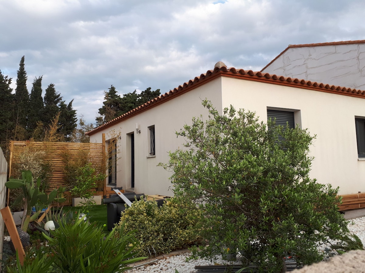 The exterior of a modern single-story villa is shown, featuring a sloped tiled roof. Lush greenery surrounds the home, including shrubs and small trees, with a wooden privacy fence partially visible in the background. The atmosphere is calm and inviting.