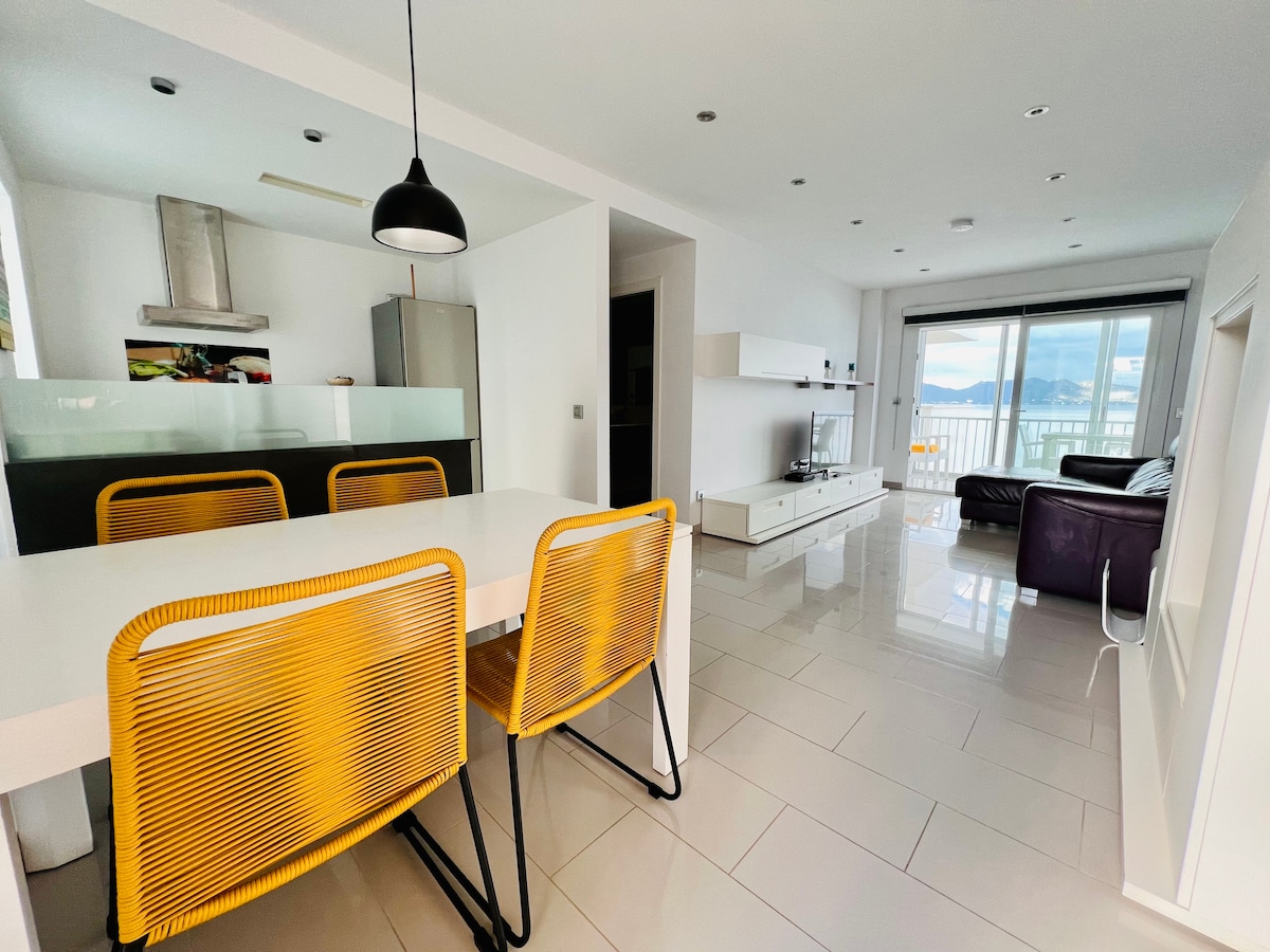 A modern open-plan living area is observed, featuring a white dining table paired with two vibrant yellow chairs. A sleek kitchen is visible in the background, complemented by a large window showcasing views of the beach. A comfortable seating area includes a dark leather sofa and a light-colored entertainment unit.
