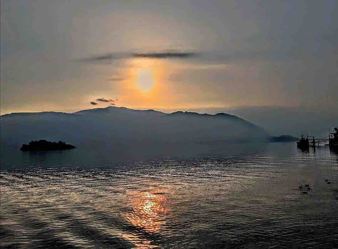 A serene lake view at sunset, with soft waves reflecting golden light. Silhouetted mountains create a tranquil backdrop, while islands are faintly visible in the distance. The scene is calm, inviting contemplation of nature's beauty.
