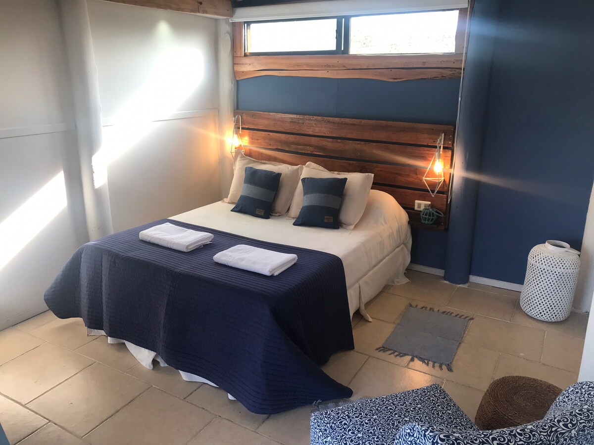 A serene sleeping area is defined by a spacious bed covered with a dark blue blanket. Two decorative pillows rest on the bed against a wooden headboard. Natural light enters through a nearby window, illuminating the light-colored tiled floor and cozy seating area.